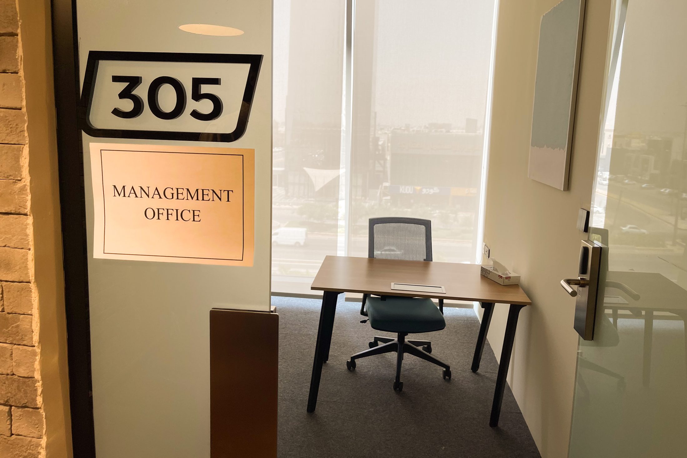 The open door of a small office, a printed sheet reading Management Office taped under the room number, an empty desk and chair inside