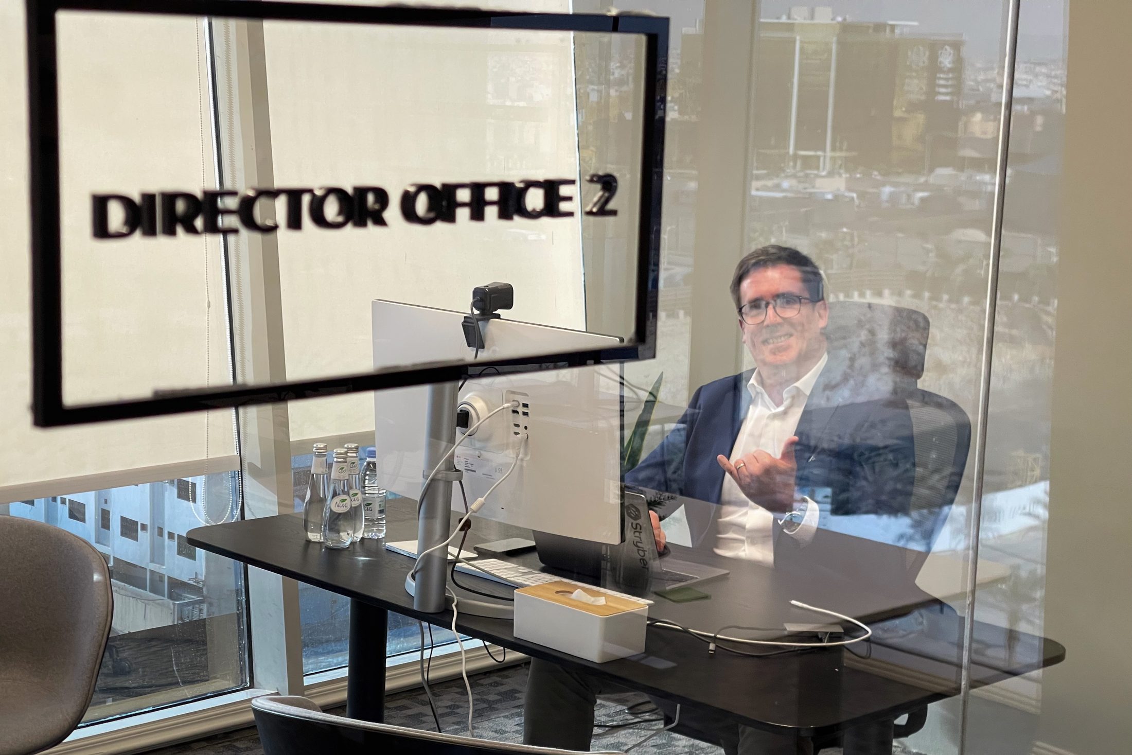 Through a glass wall lettered Director Office 2, a man in a dark jacket sits at a desk by a window, smiling and gesturing, a monitor and water bottles on the desk