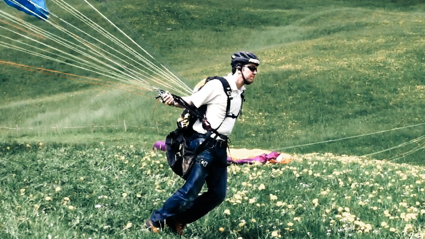 A man in a helmet and sunglasses runs down a meadow full of dandelions, pulling the lines of a paraglider behind him