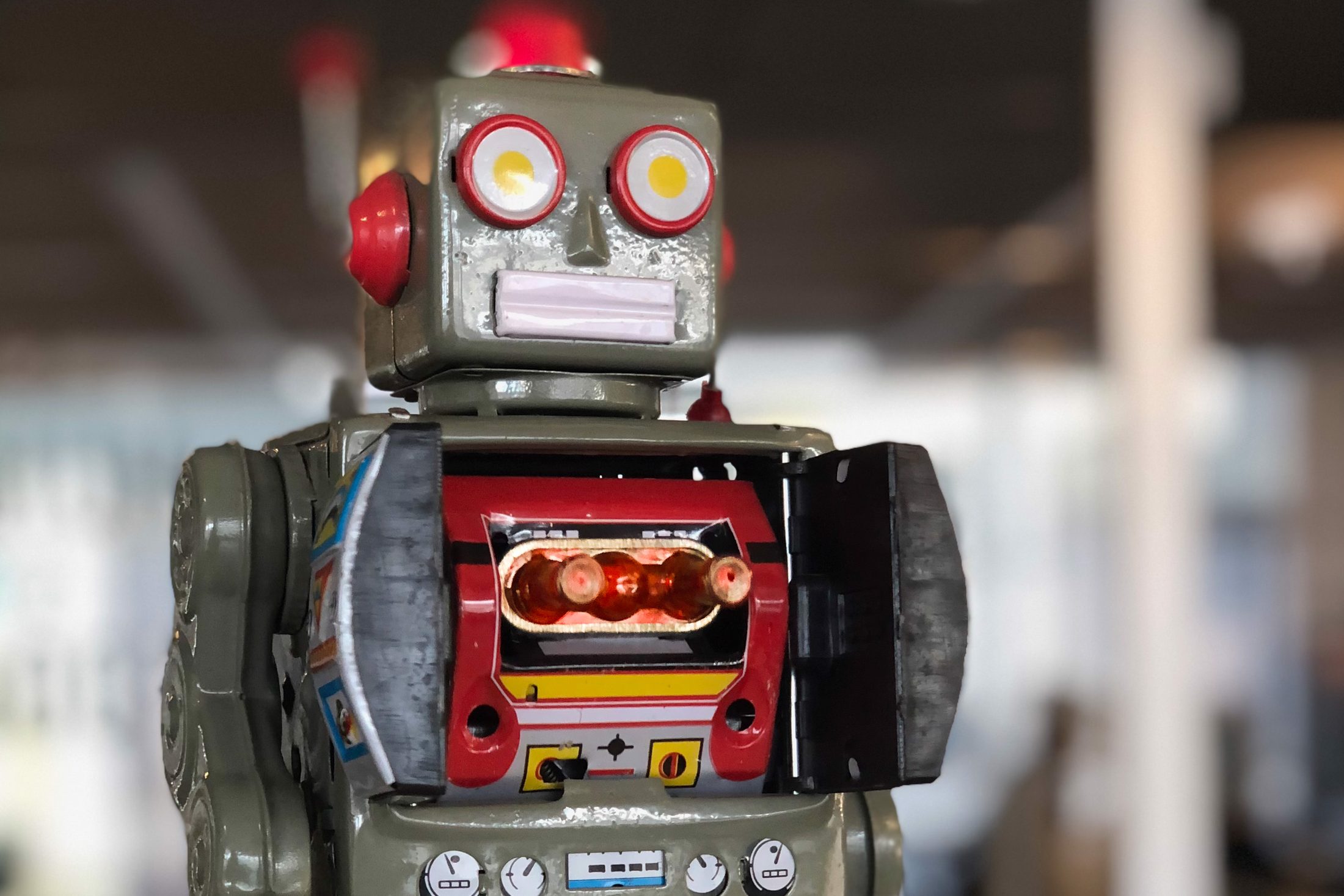 A vintage tin toy robot with round red-rimmed eyes and an open chest door showing glowing red pistons, an office blurred behind it