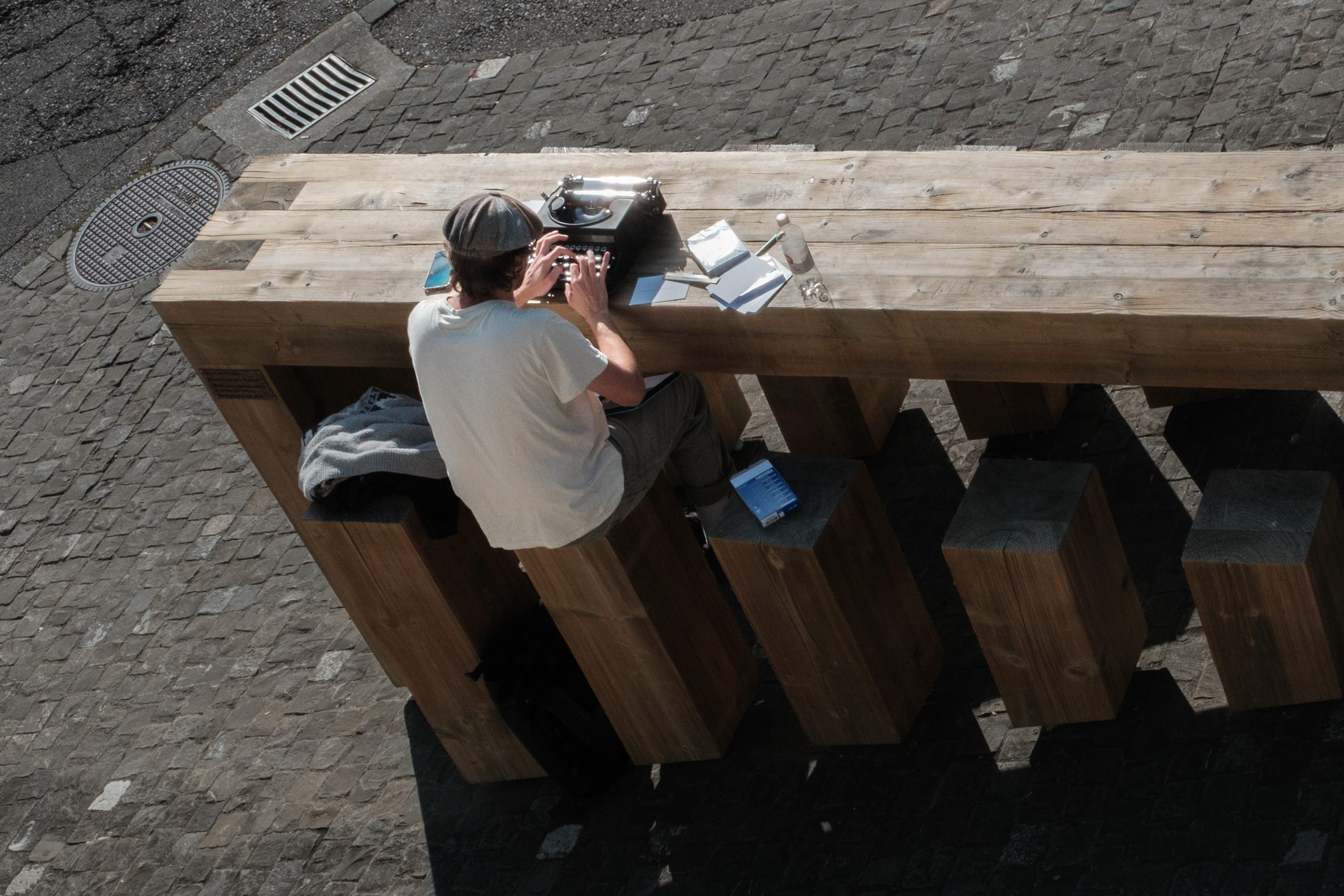 A man at a long wooden table in a cobbled square, typing on a typewriter, seen from above